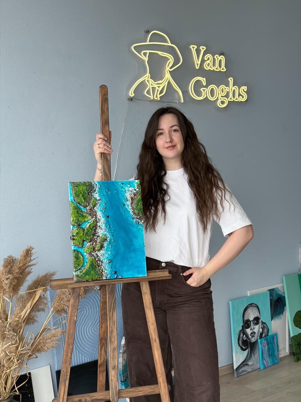 Засновниця Van Gogh's Studio Катерина стоїть біля картини з епоксидної смоли з зображенням островів під неоновою вивіскою студії